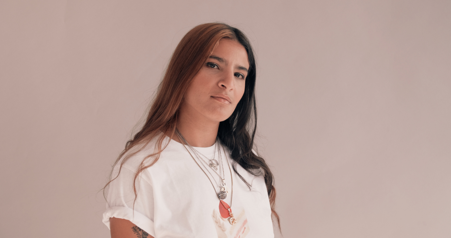 A person of colour in a white shirt with necklaces and coloured hair against a neutral background